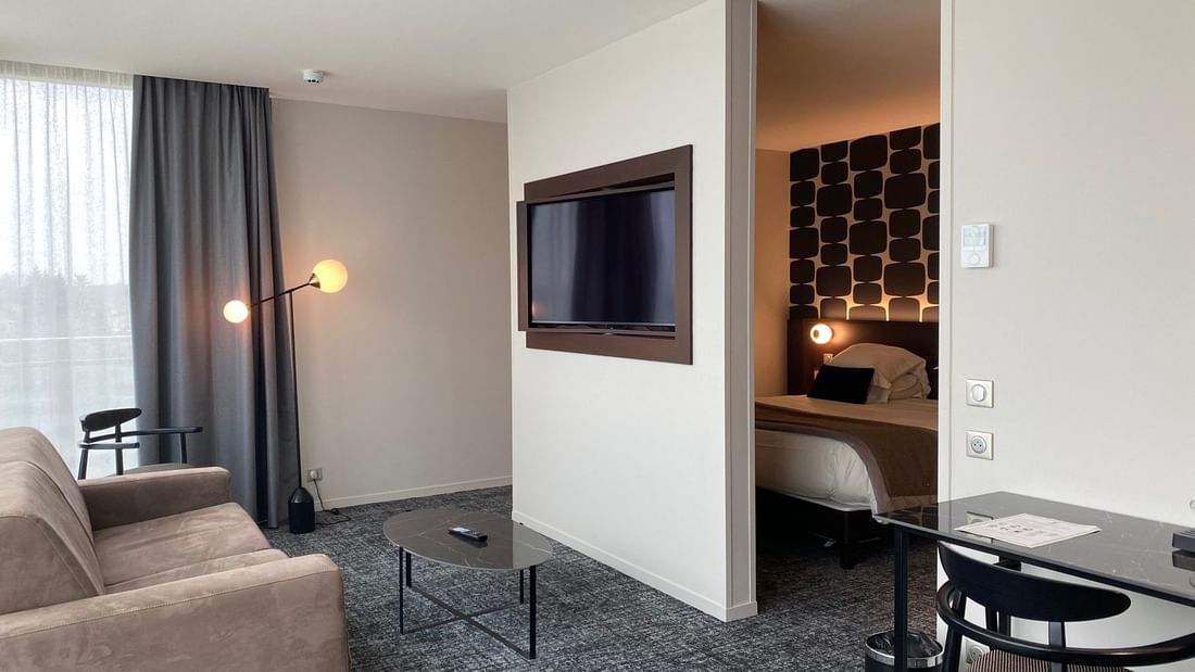 Chambre d'hôtel avec canapé, table basse, bureau, TV murale, lit dans une pièce adjacente.