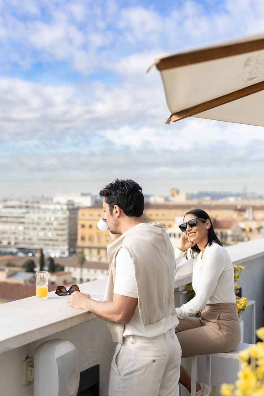 A couple enjoys drinks on a rooftop terrace at The Independent, with a scenic city skyline under a cloudy sky