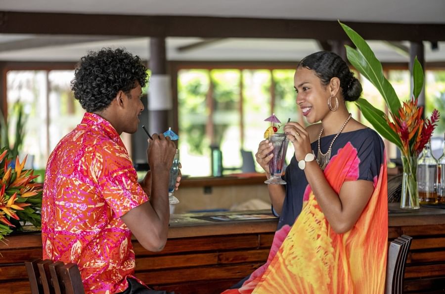 Couple in vibrant attire enjoying drinks by a wooden bar in Tambua Restaurant at Tambua Sands Beach Resort