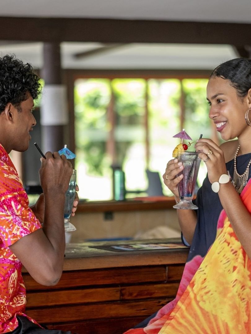 Couple in vibrant attire enjoying drinks by a wooden bar in Tambua Restaurant at Tambua Sands Beach Resort