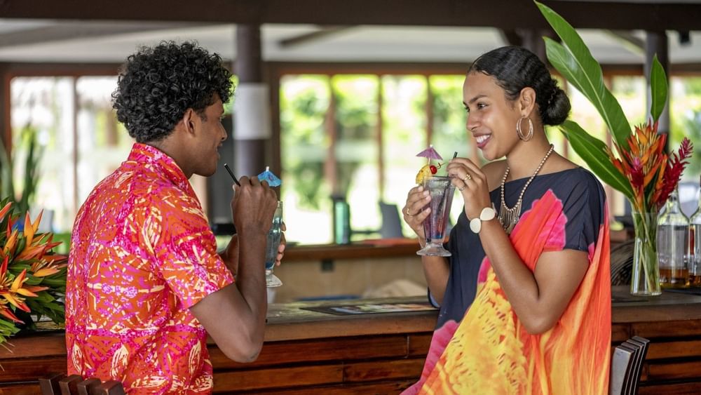 Couple in vibrant attire enjoying drinks by a wooden bar in Tambua Restaurant at Tambua Sands Beach Resort