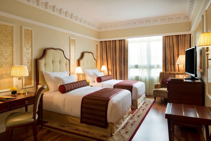 Twin beds by a wooden desk under bright lamps near a large window and television in Superior Room at Warwick Doha Hotel