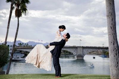 Groom lifts bride in lace gown on waterfront lawn with bridge and palm trees at London Bridge Resort