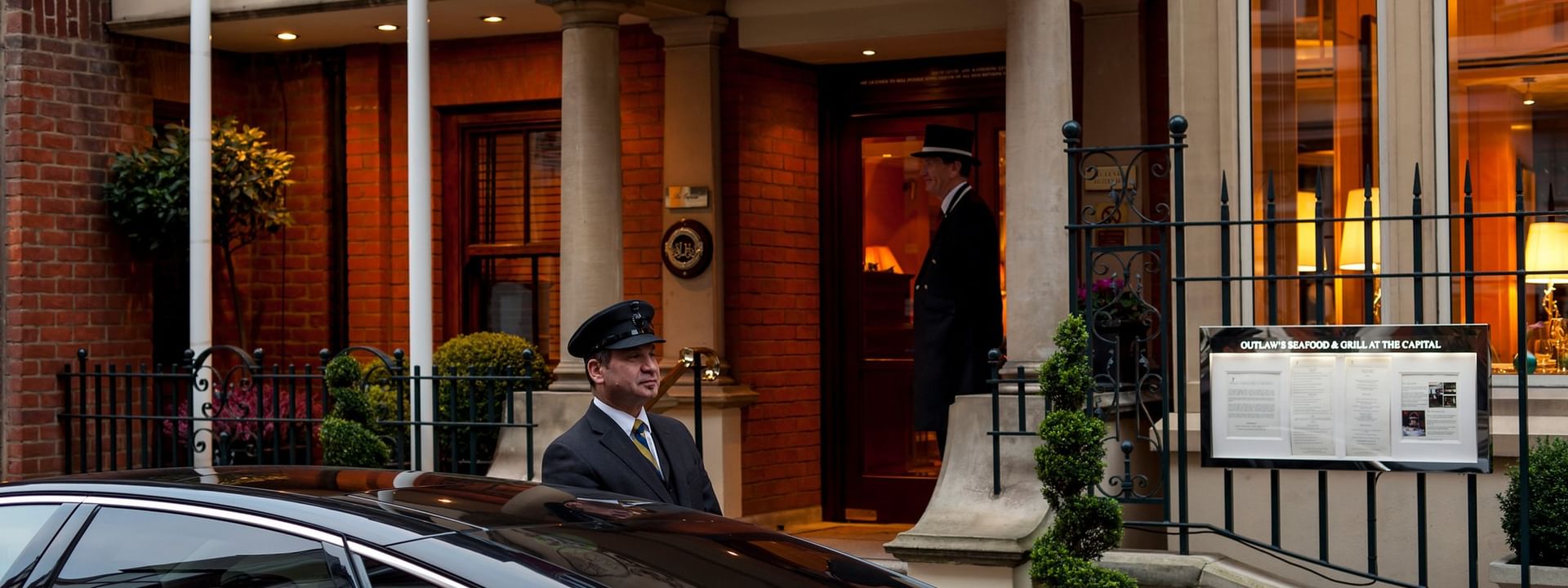 Doorman in a formal uniform standing by a black car near the entrance of The Capital Hotel, Apartments and Townhouse