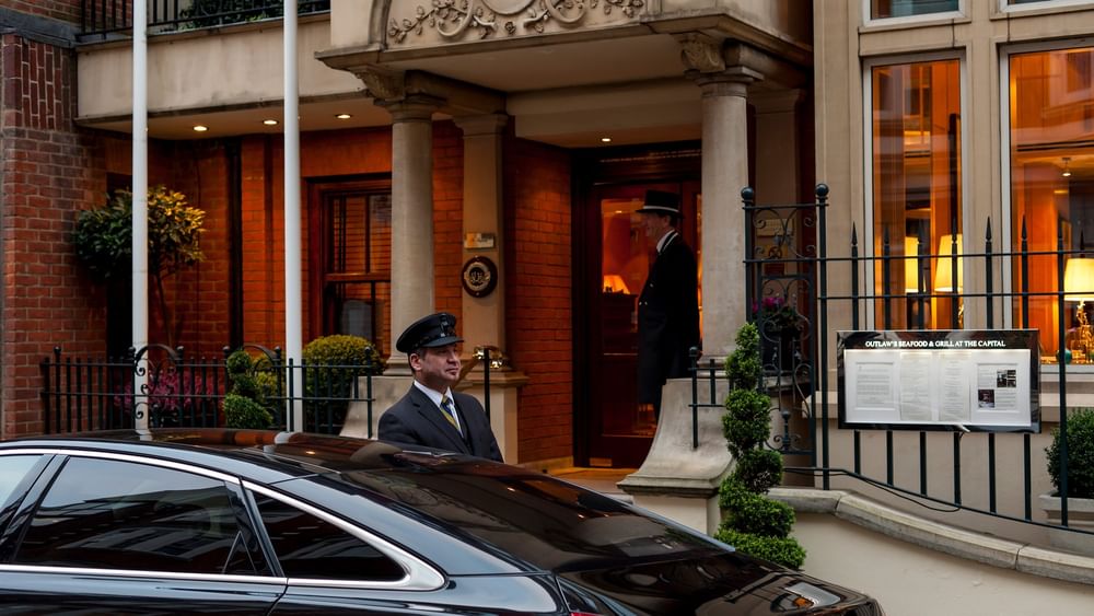 Doorman in a formal uniform standing by a black car near the entrance of The Capital Hotel, Apartments and Townhouse