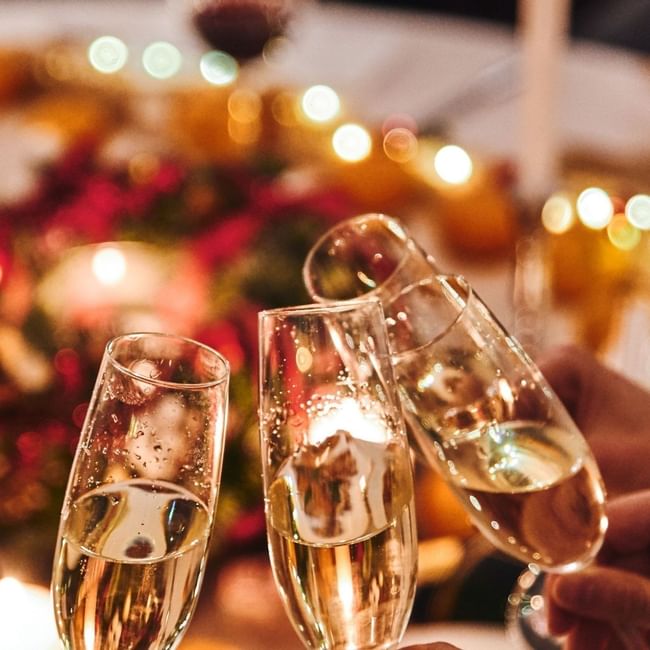 Three champagne flutes toasting over a festive Christmas Day Lunch table with warm lights and holiday decorations.