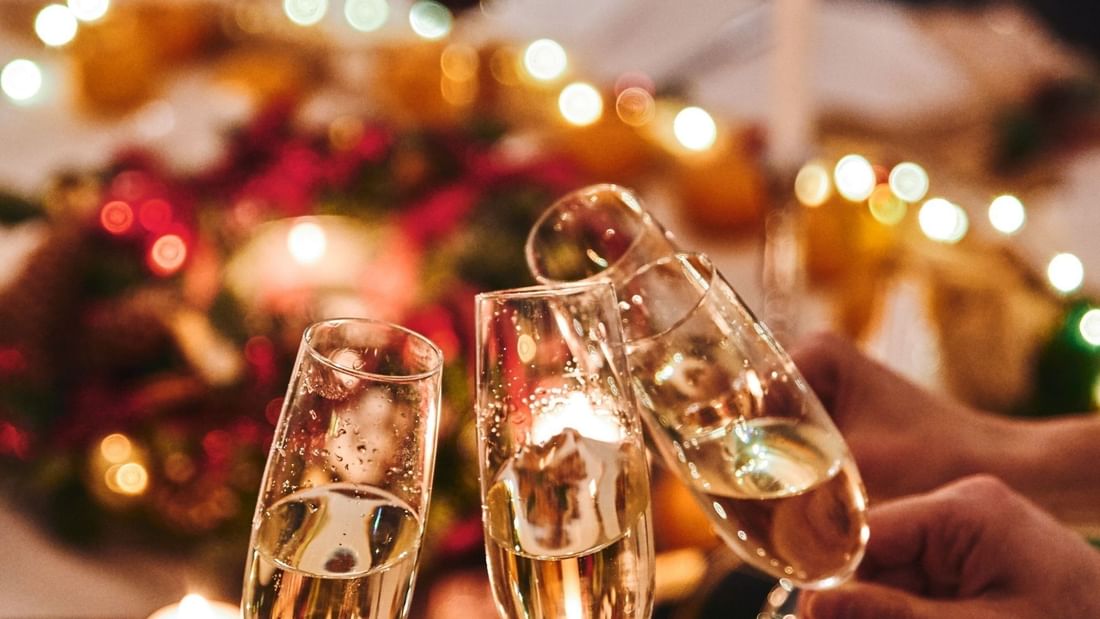 Three champagne flutes toasting over a festive Christmas Day Lunch table with warm lights and holiday decorations.