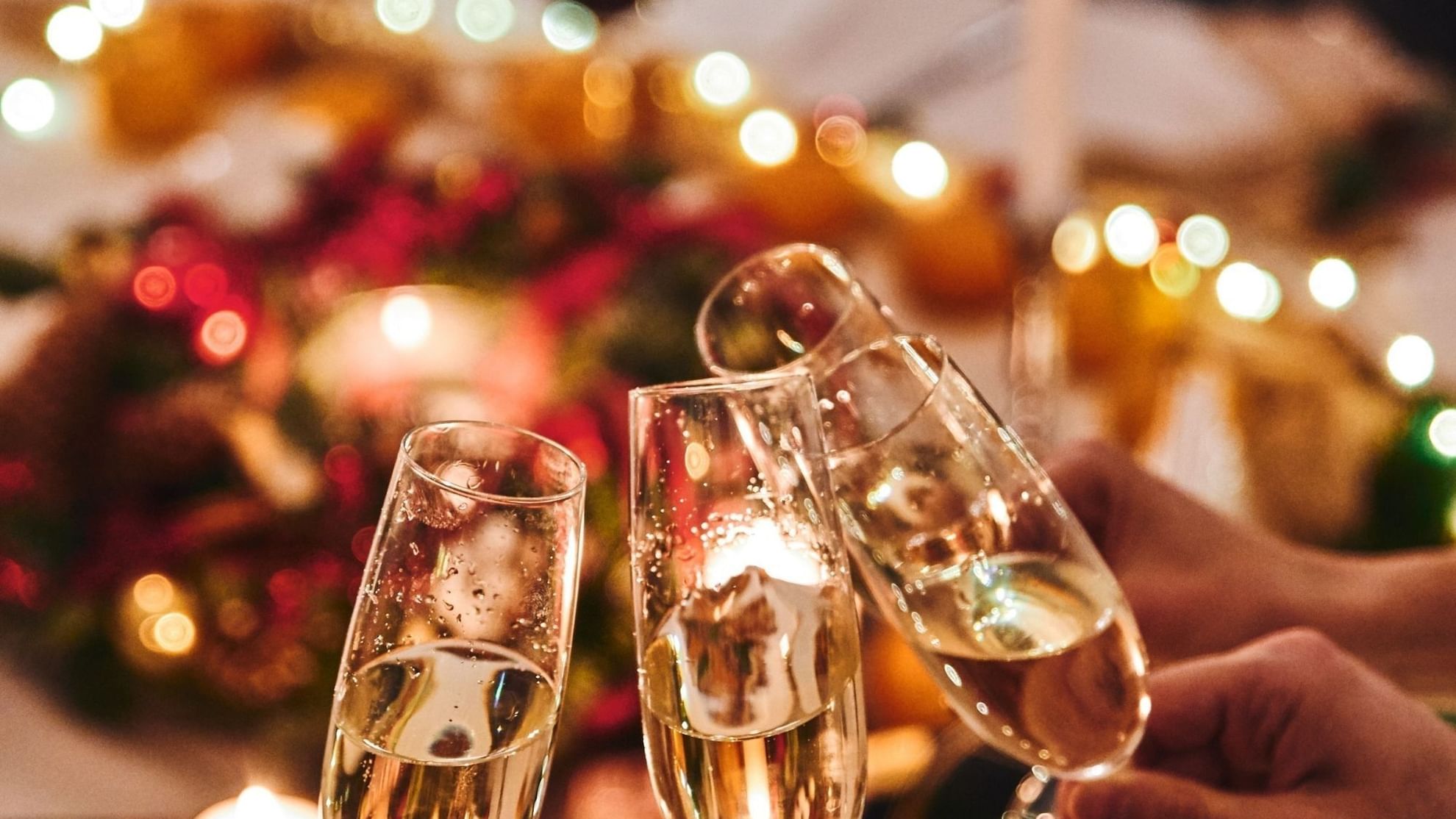 Three champagne flutes toasting over a festive Christmas Day Lunch table with warm lights and holiday decorations.