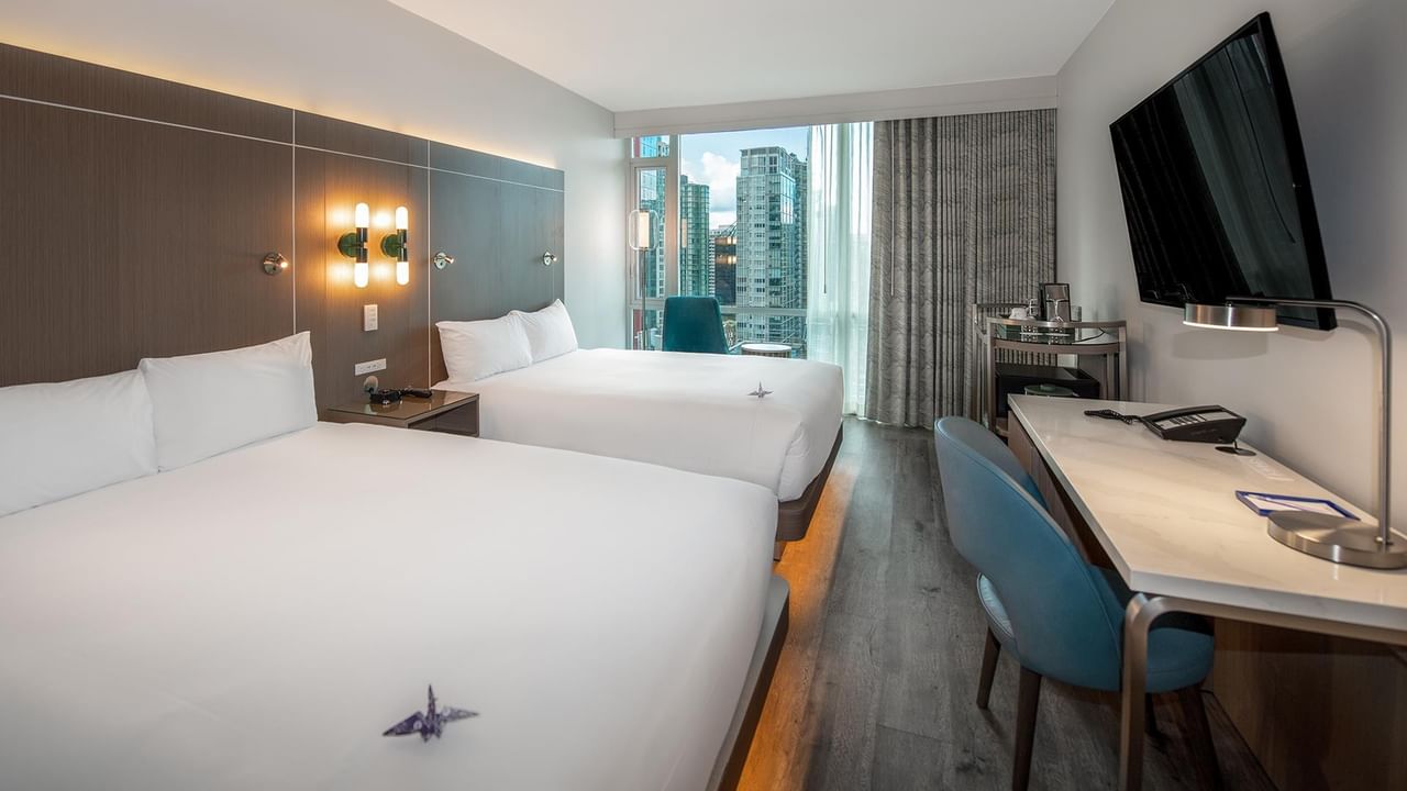 Two beds, desk, chair, and cityscape view through a large window.