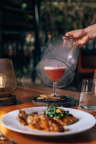 Person unveils a smoked cocktail next to a plate of food in a restaurant at El Mangroove Hotel