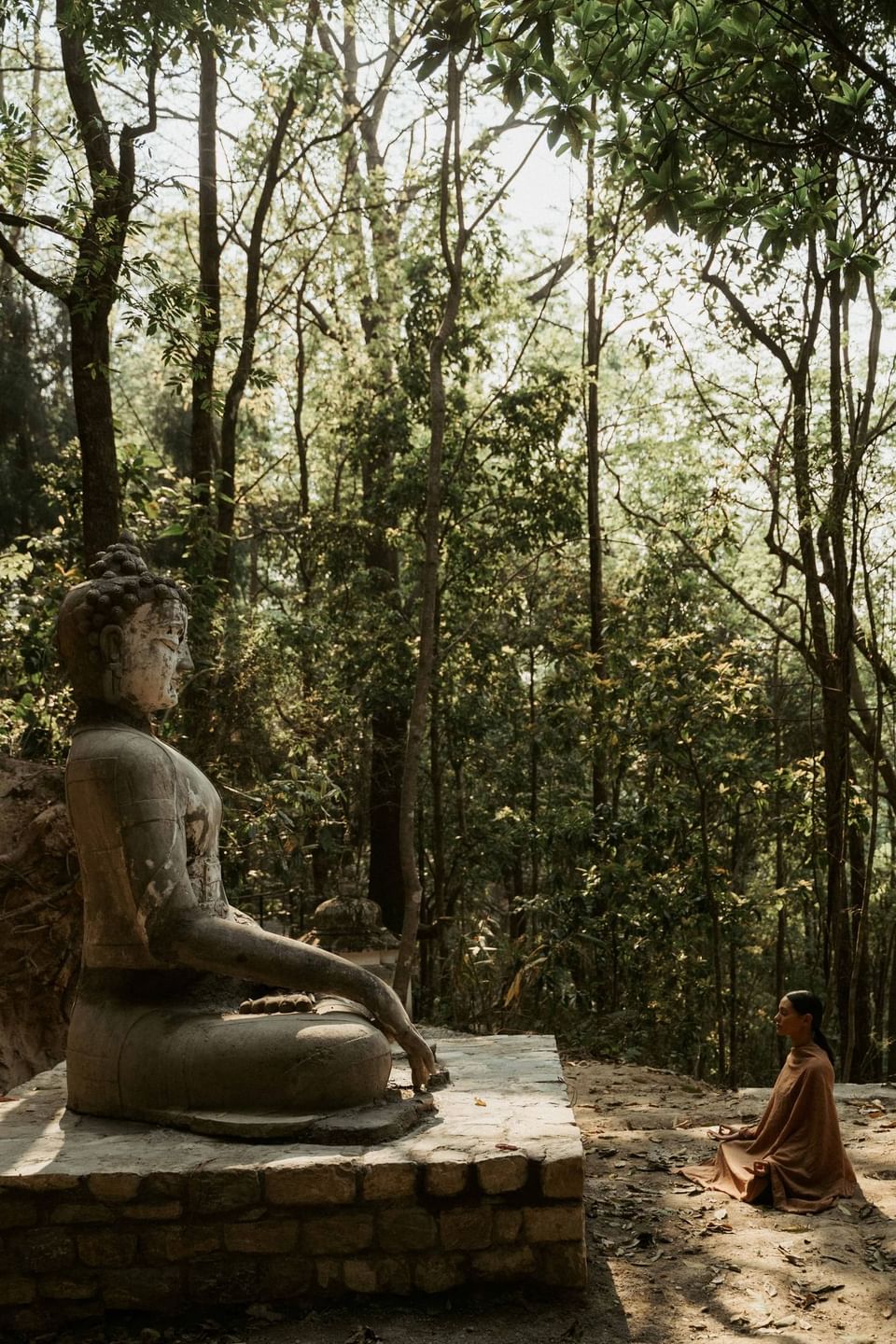 Buddhist statue on stone platform in lush forest with person meditating nearby at The Dwarika's Himalayan Wellbeing
