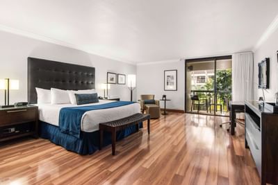 King bed with blue throw beside an armchair and glass door opening to a balcony in the Junior Suite at Miami Lakes Hotel