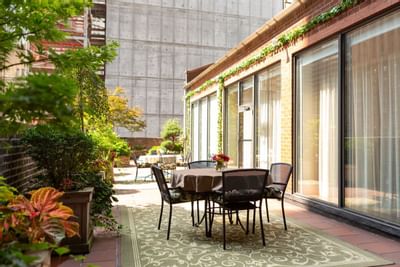 Outdoor patio with table and chairs, large plants, brick building, and glass doors with curtains.