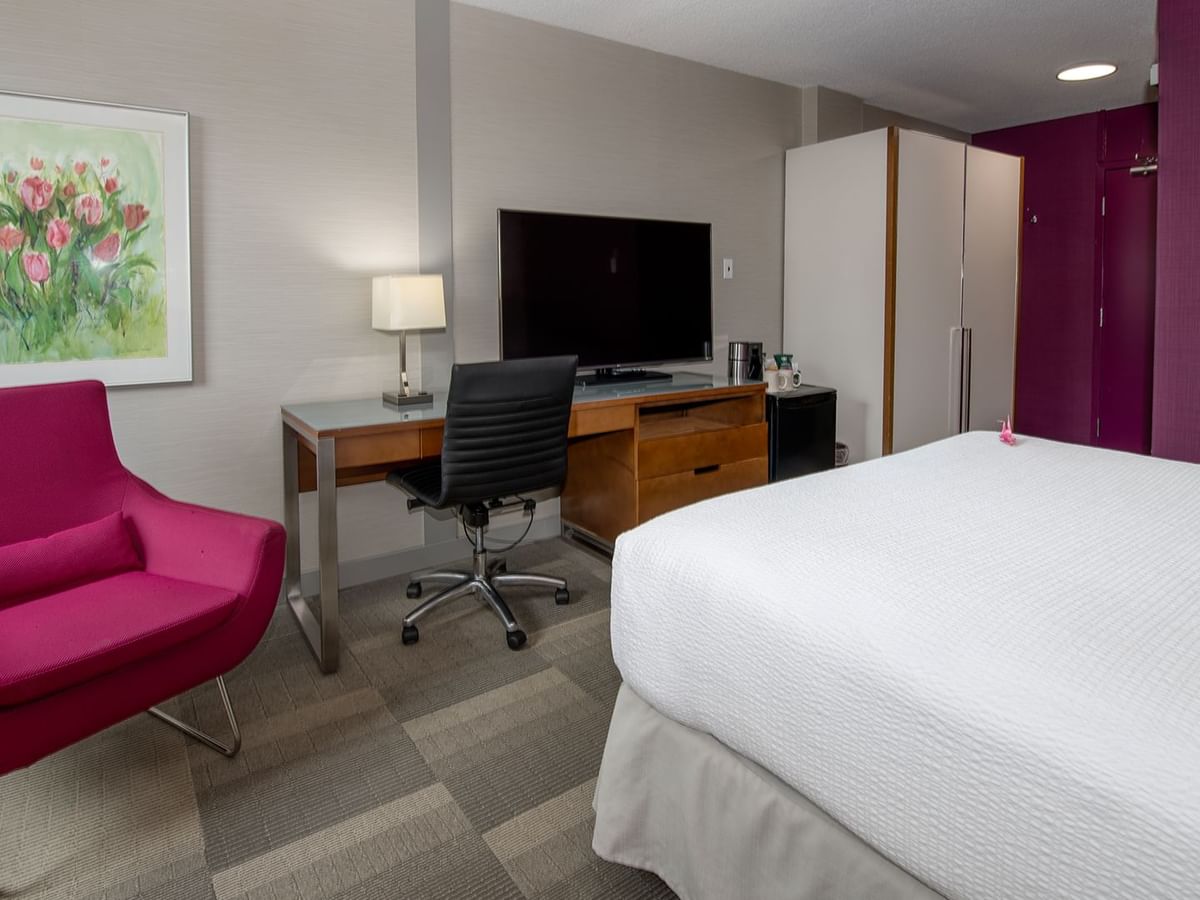 A hotel room with a white bed, desk with TV, pink chair, and a framed painting.