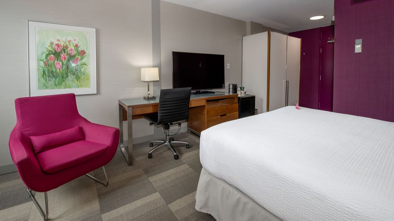 A hotel room with a white bed, desk with TV, pink chair, and a framed painting.