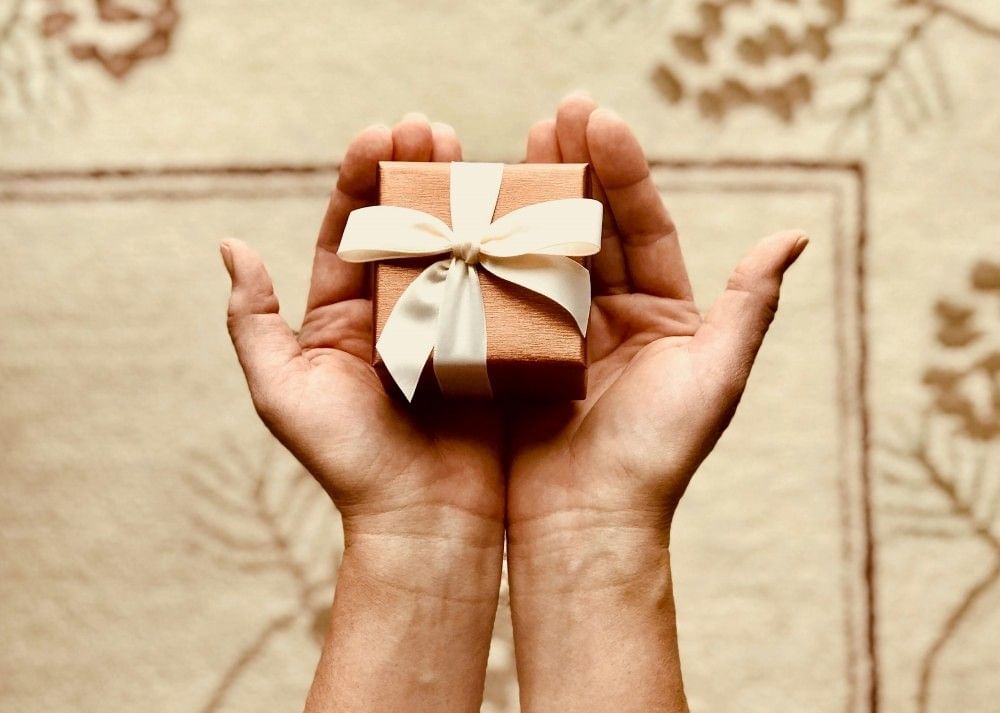 Outstretched hands holding a small parcel wrapped in brown paper and beige ribbon.