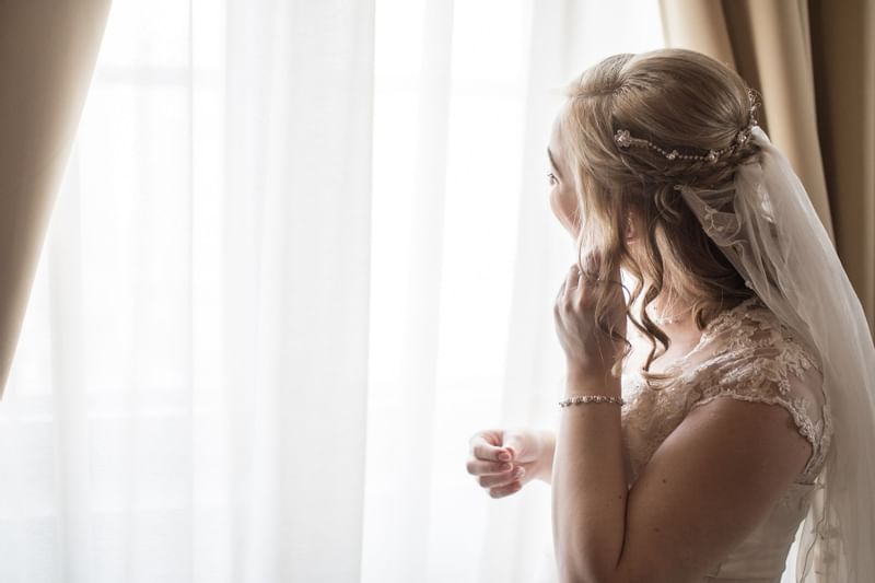 Bride in wedding gown and veil looking out of window, touching her hair.
