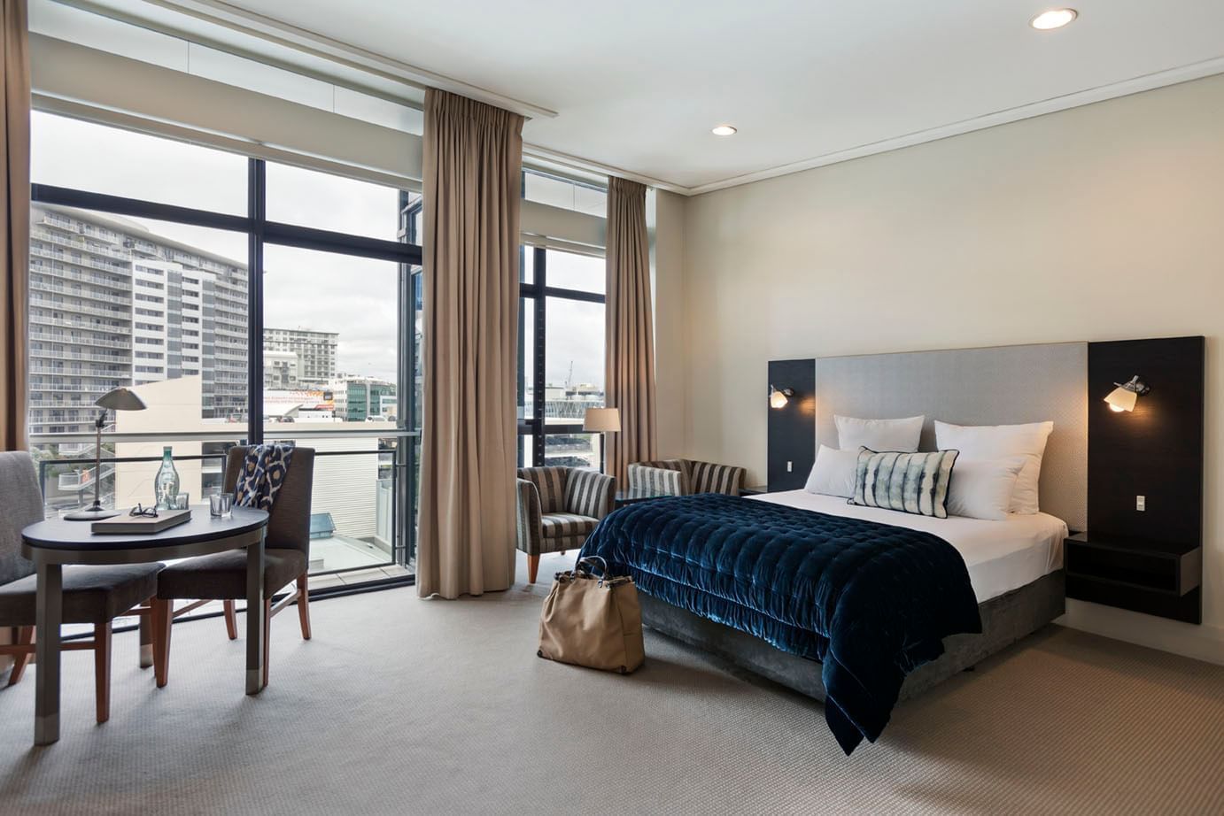 Bed with seating and dining table by the window overlooking buildings in City View Studio at Sebel Auckland Viaduct Harbour