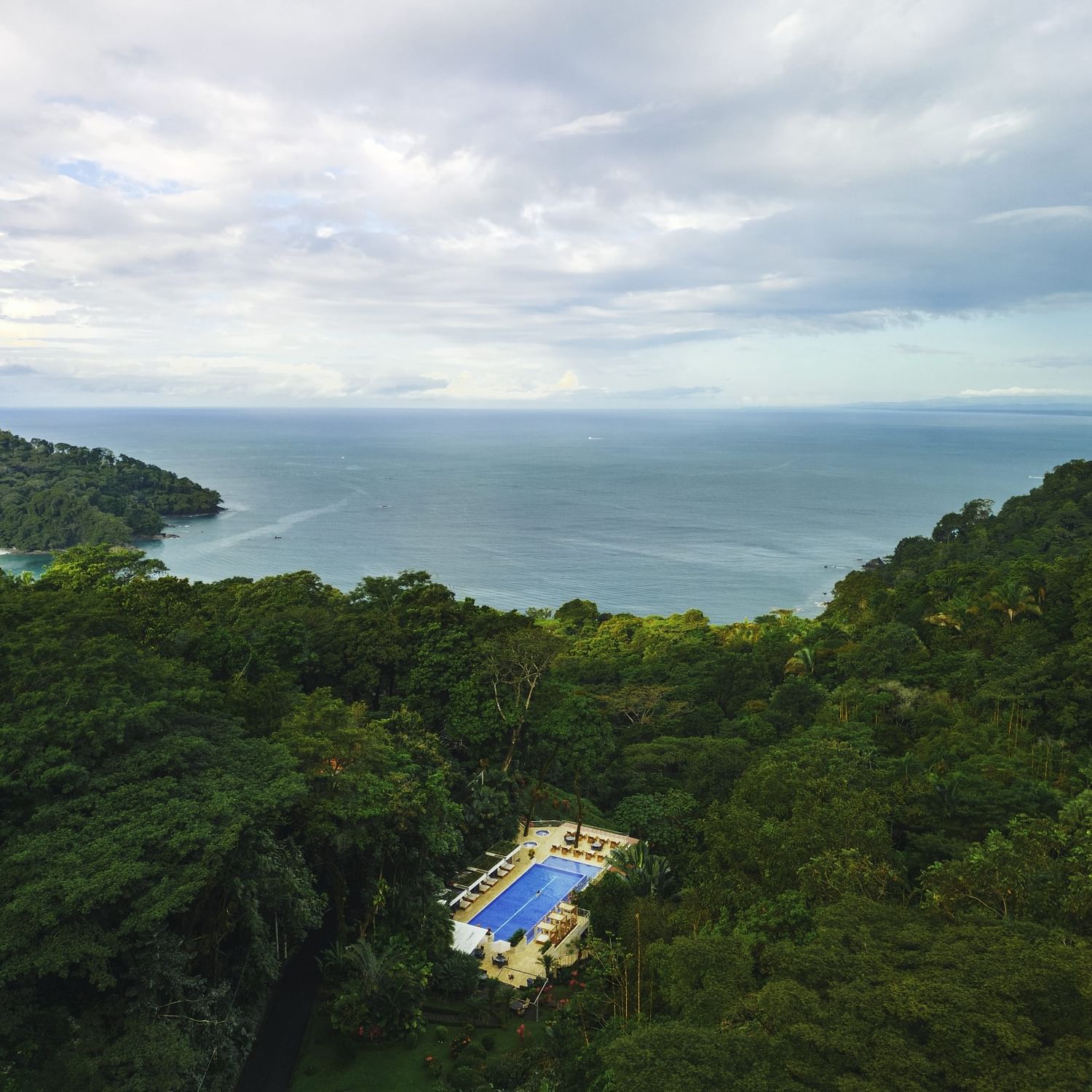 Aerial view of a resort pool surrounded by lush rainforest with ocean and coastal hills under a cloudy sky in Costa Rica