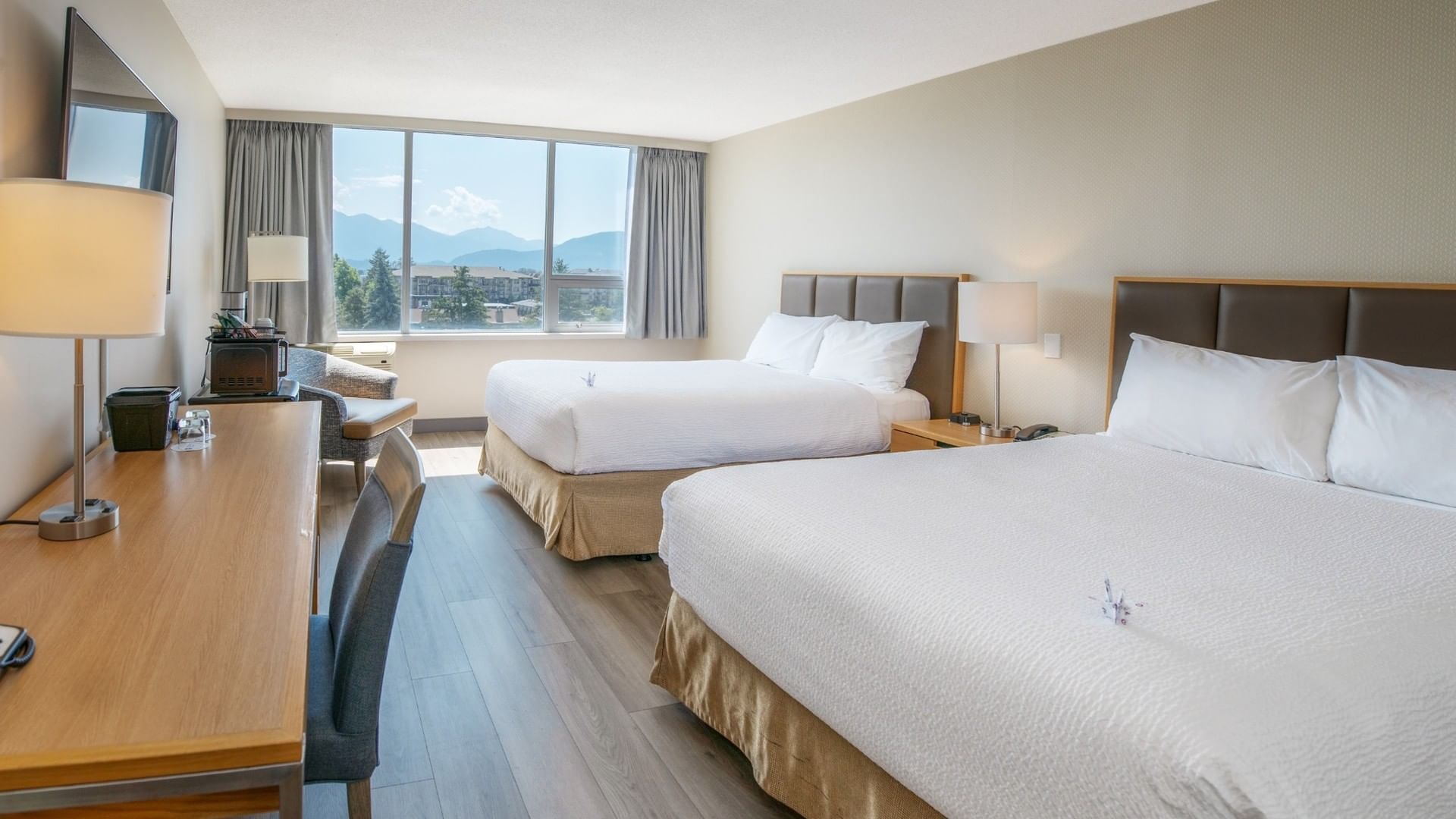 Two hotel beds, desk with lamp, and chair in room with large window showing mountain view.
