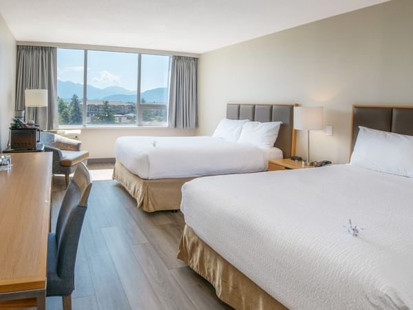 A hotel room with two beds, work desk with lamp, chair, TV, and large window with pond view.