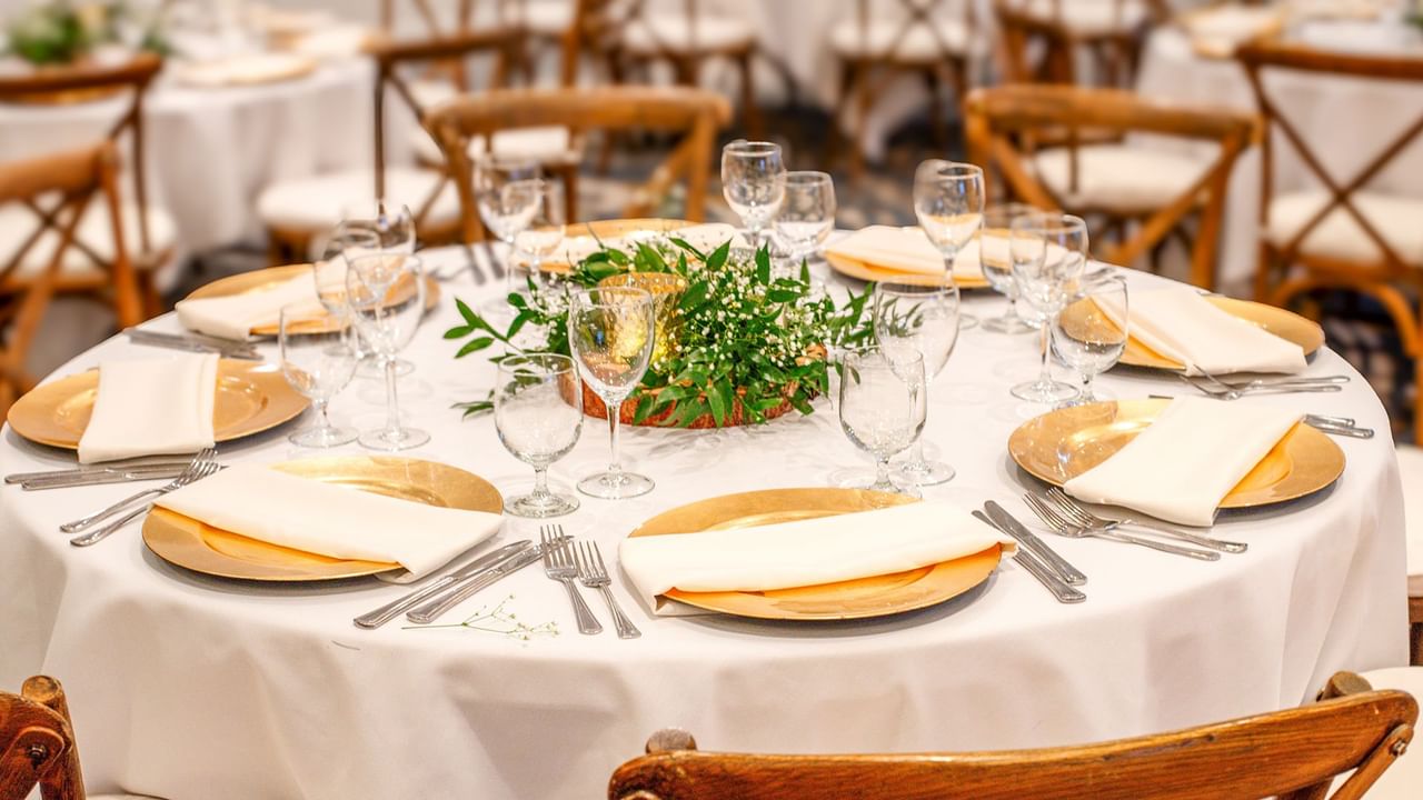A dining table set with white tablecloth, gold plates, napkins, silverware, wine glasses, and a flower centerpiece.