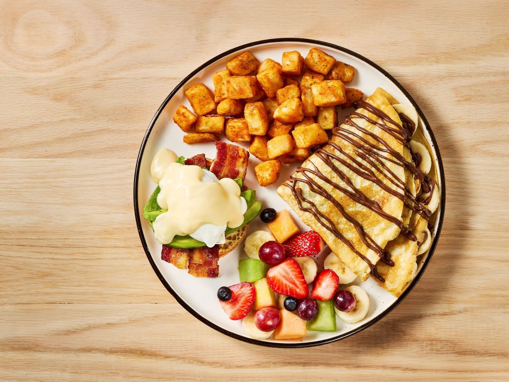 Plate with a waffle, bacon, hollandaise sauce, fruit salad, and hash browns.