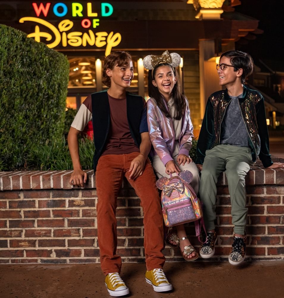 Three teenagers sit on a brick half-wall in front of the colorful sign for World of Disney.