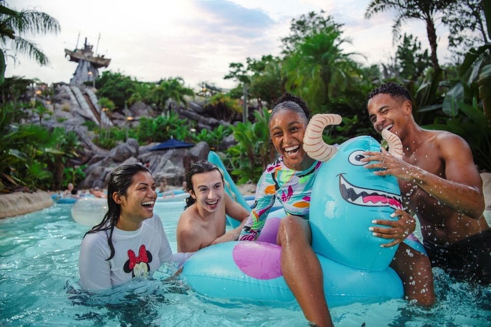 Two men and two women in bathing suits laugh and surround a blue Monsters Inc. themed inner tube, with the typhoon lagoon ship teetering on a mountain in the distance.