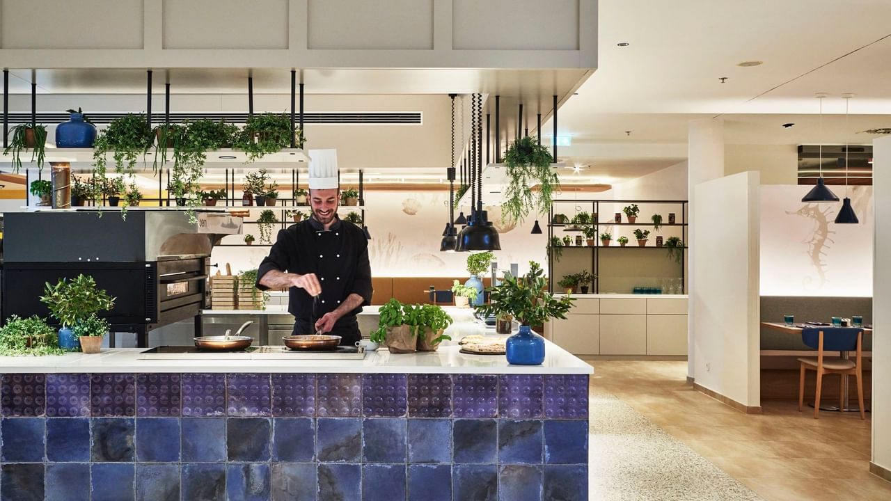 Chef cooking in a kitchen with hanging plants and blue tile decor at Falkensteiner Hotel & Spa Carinzia