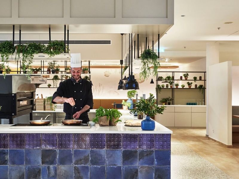 Chef cooking in a kitchen with hanging plants and blue tile decor at Falkensteiner Hotel & Spa Carinzia