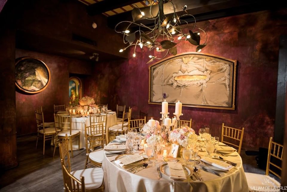 A dining room with round tables, gold chairs, lit candles, and decorative paintings on the wall.