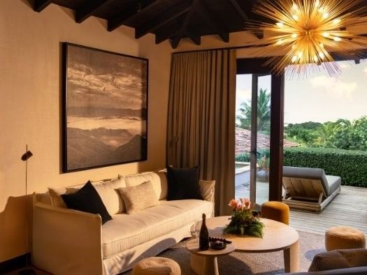 Modern living room with cream sofa, starburst chandelier, and outdoor terrace with lounge chairs and lush greenery.