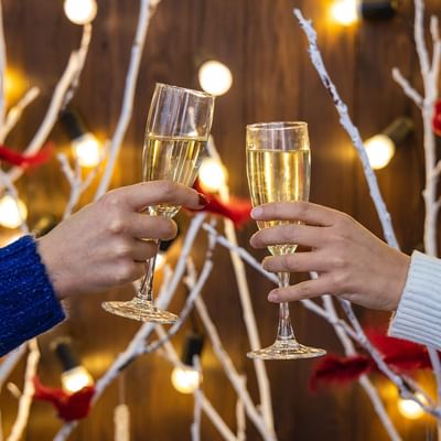 Zwei Hände stoßen mit Sektgläsern an, vor einem Hintergrund mit Lichtern und Zweigen.
