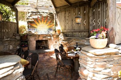 Outdoor dining area with brick fireplace, wooden table, stone walls, and patio chairs.
