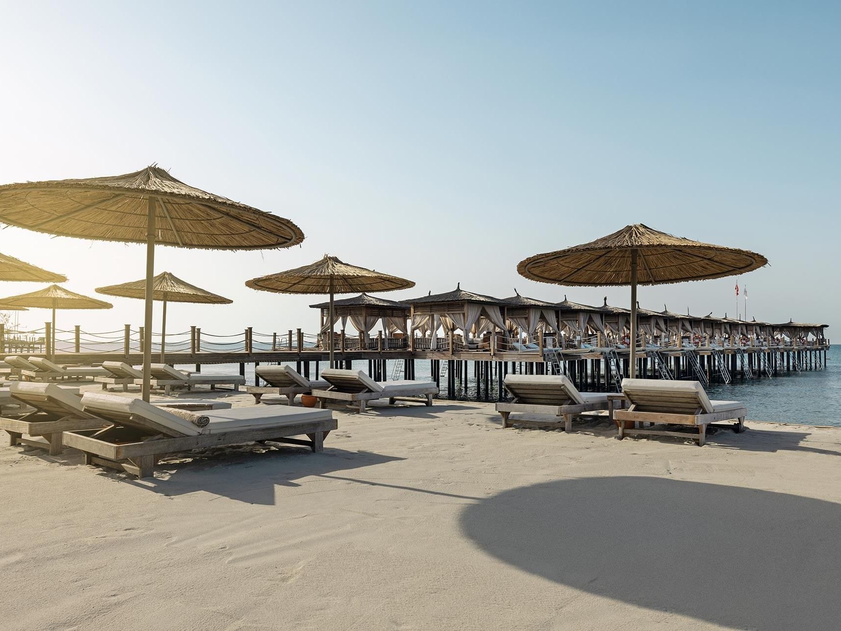 Beach with lounge chairs, umbrellas, and a pier stretching into the water under a clear sky Cullinan Belek.