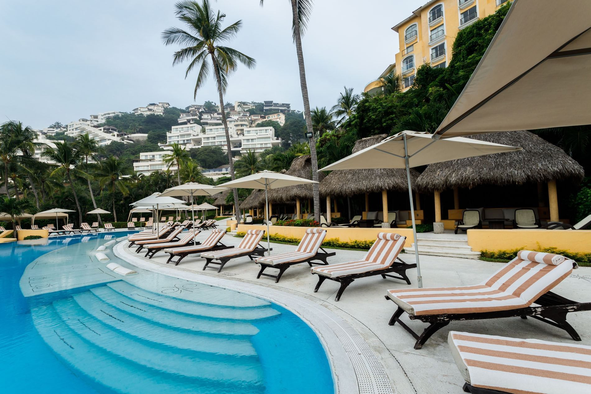 Curving pool lined with striped lounge chairs, umbrellas, and tropical palm trees at Quinta Real Acapulco
