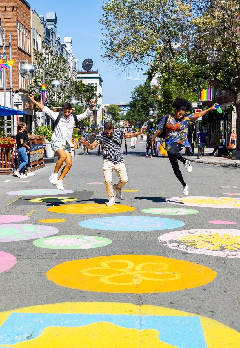 Three friends jumping joyfully on a colorful painted pedestrian street lined with shops and trees at Warwick Le Crystal Montreal