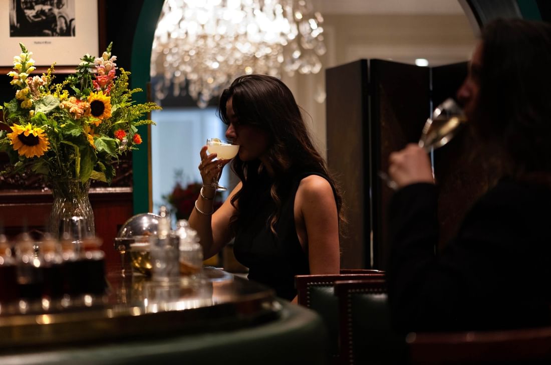 Woman sipping a cocktail by a large flower vase, under a sparkling chandelier in Duke's Bar at Hotel Westminster Paris