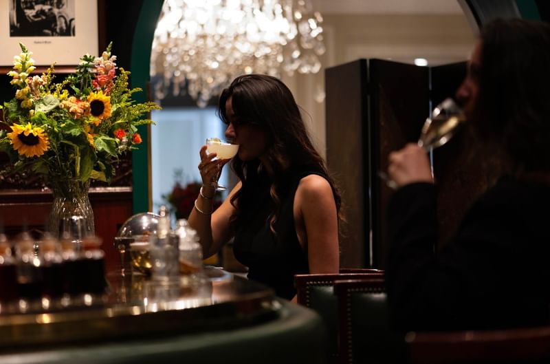 Une femme déguste un cocktail au bar élégant avec lustre et bouquet de tournesols à Hotel Westminster Warwick Paris