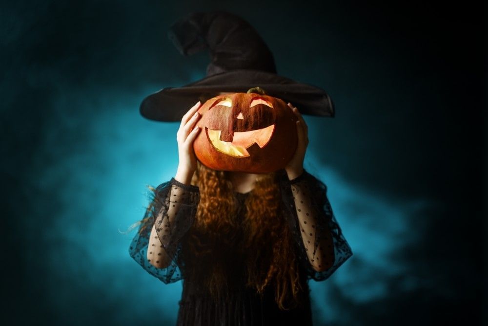 Celebrating Halloween in Orlando, a girl in a witch costume stands in front of a hazy blue and black background, holding a glowing, smiling jack-o-lantern in front of her face.