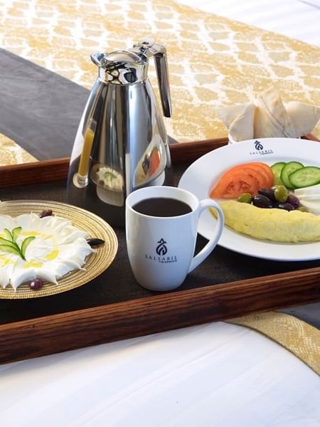 Breakfast tray on a bed with a silver coffee pot, orange juice, & a cream dessert in the Family Suite at Warwick Hotel Jeddah