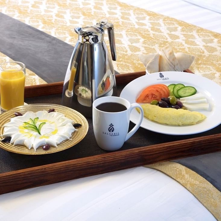 Breakfast tray on a bed with a silver coffee pot, orange juice, & a cream dessert in the Family Suite at Warwick Hotel Jeddah