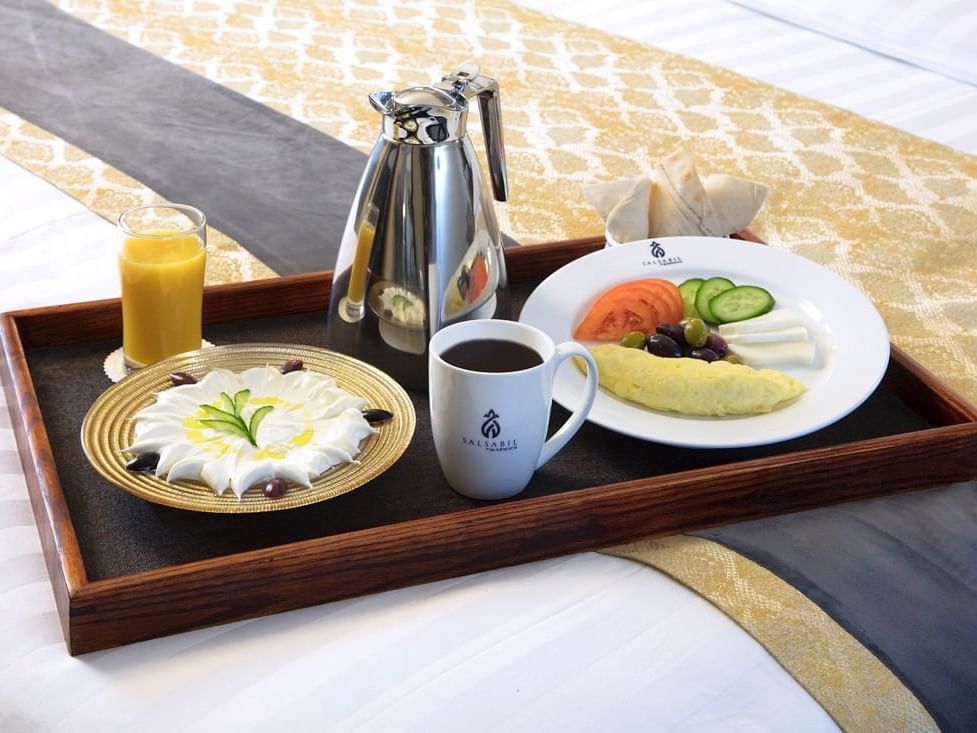Breakfast tray on a bed with a silver coffee pot, orange juice, & a cream dessert in the Family Suite at Warwick Hotel Jeddah