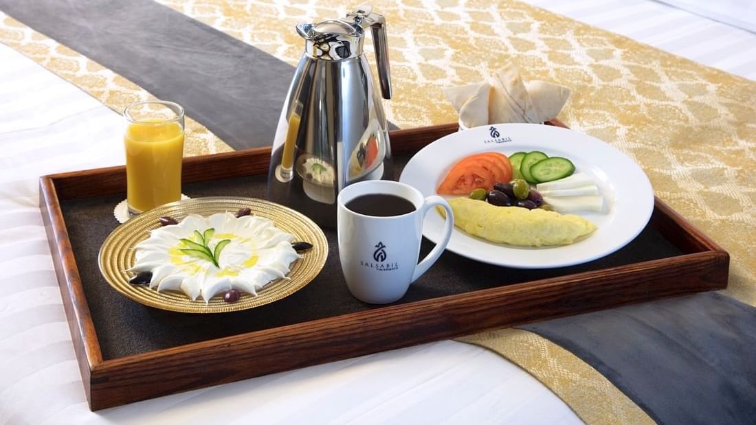 Breakfast tray on a bed with a silver coffee pot, orange juice, & a cream dessert in the Family Suite at Warwick Hotel Jeddah