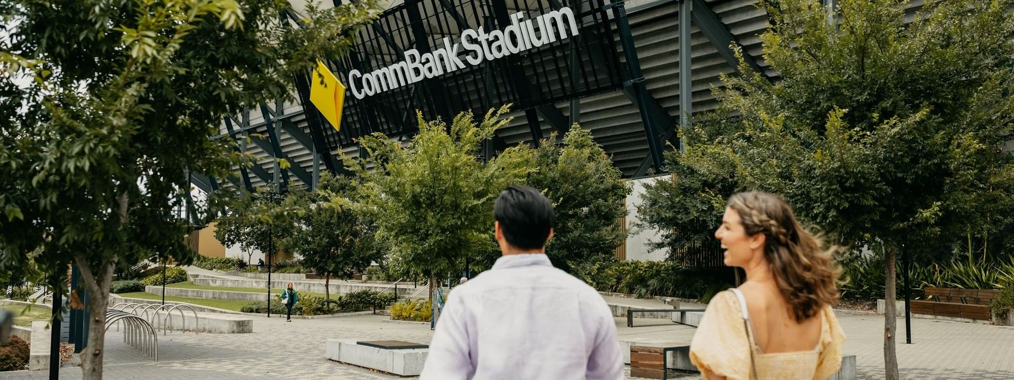 Two people walk towards CommBank Stadium with trees and seating areas nearby.