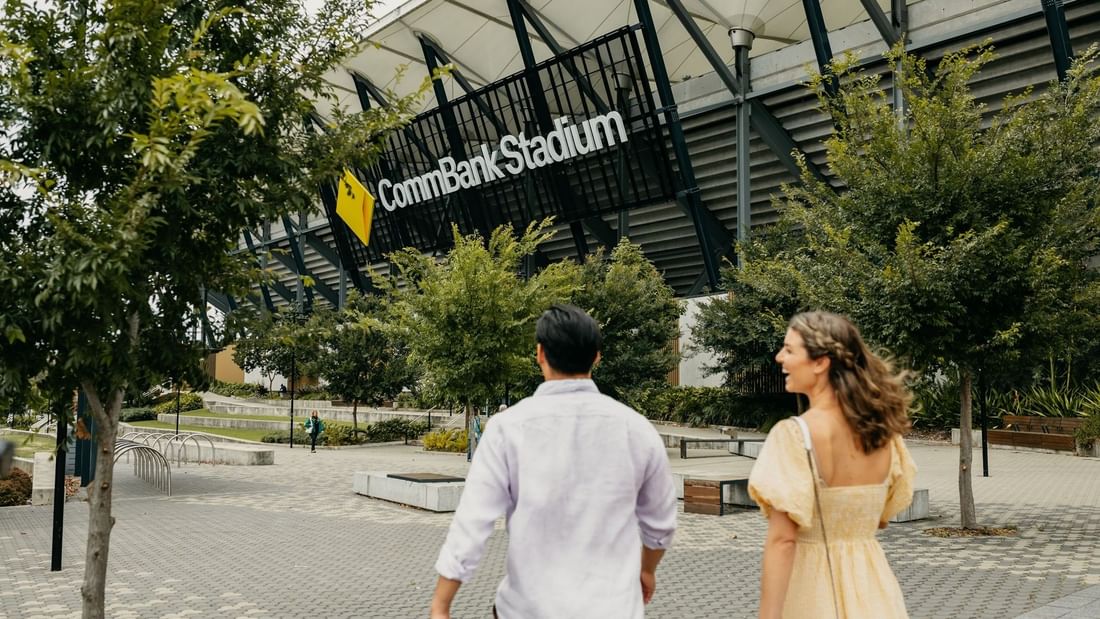 Couple walking towards the entrance of the CommBank Stadium with trees and benches in the foreground.
