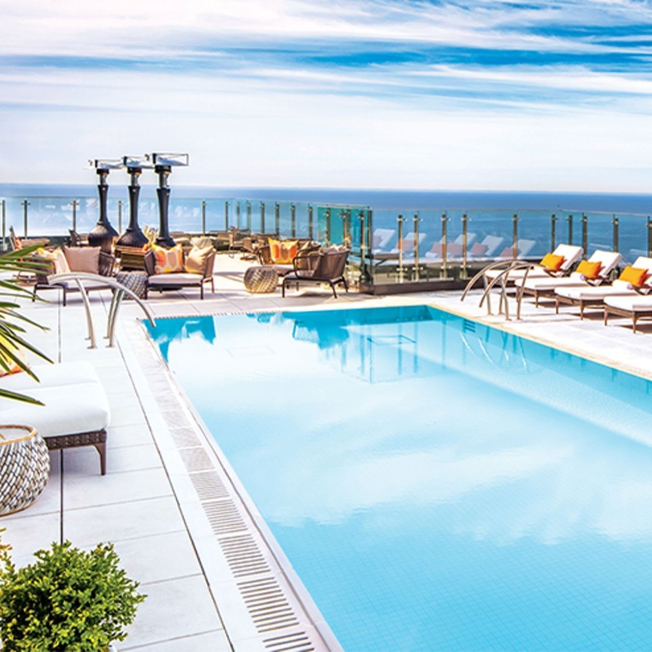 Rooftop pool by lounge chairs and fire heaters, surrounded by a glass railing overlooking the ocean at Hotel X Toronto