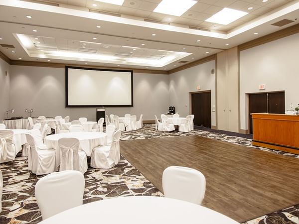 Large conference room with tables, chairs, wooden floor, and projector screen.