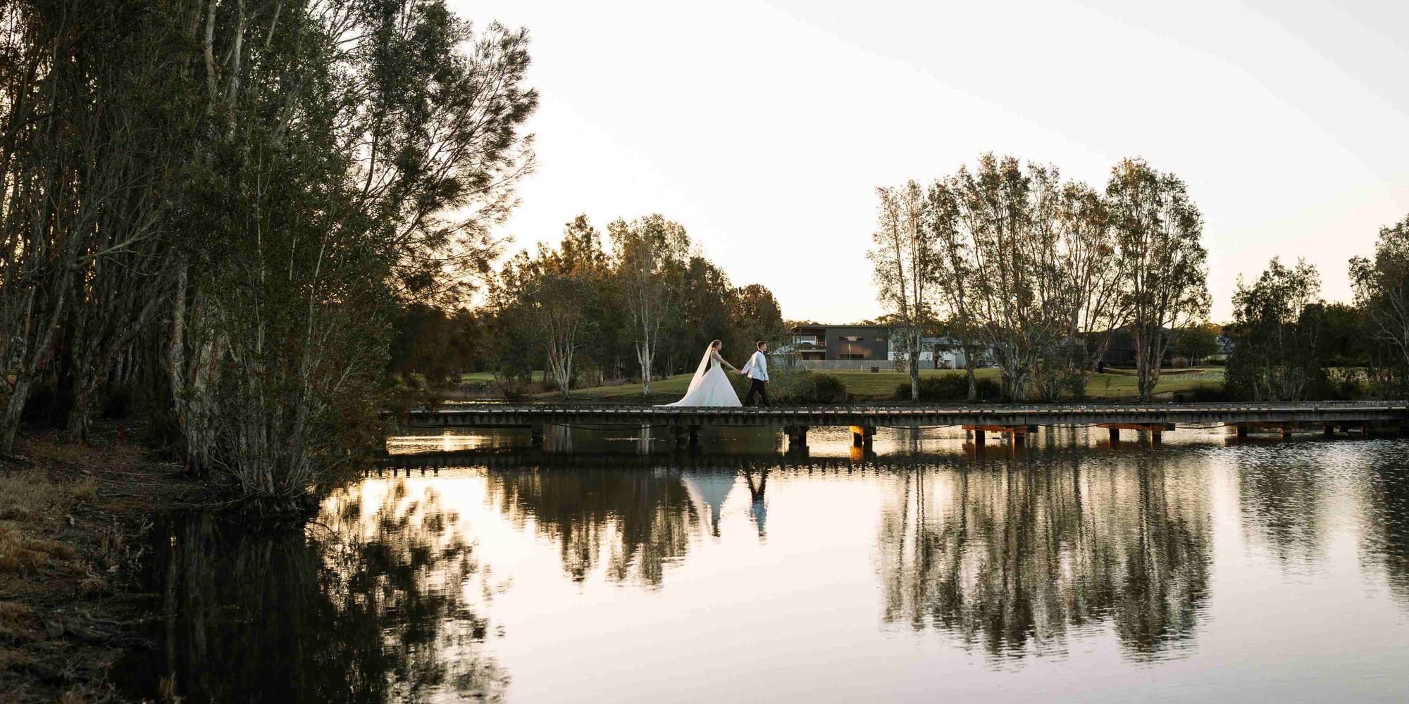 Wedded couple holding hands on a jetty by a lake at sunset near Mercure Kooindah Waters